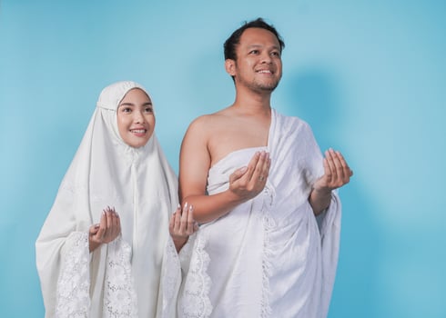 Smiling young Asian Muslim couple wearing ihram and prayer gown with praying hand gesture over isolated by blue background. Hajj concept. photo