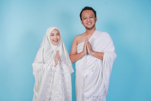 Lovely young Asian Muslim couple in ihram and prayer gown doing traditional greeting gesture, isolated by blue background. Hajj concept. photo