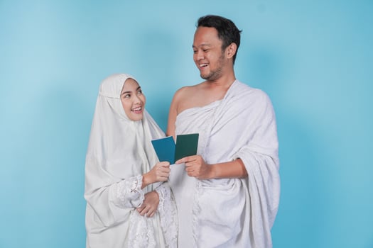 Young Asian Muslim couple in ihram and prayer gown holding a passport while looking at each other, isolated by blue background. Hajj concept. photo