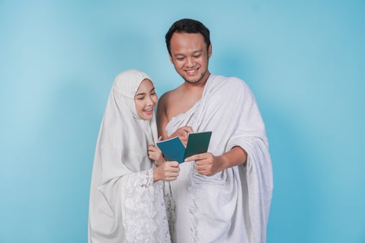 Young Asian Muslim couple in ihram and prayer gown looking at each other passport, isolated by blue background. Hajj concept. photo