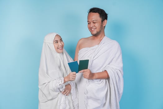 Young Asian Muslim couple in ihram and prayer gown holding a passport while looking at each other, isolated by blue background. Hajj concept. photo