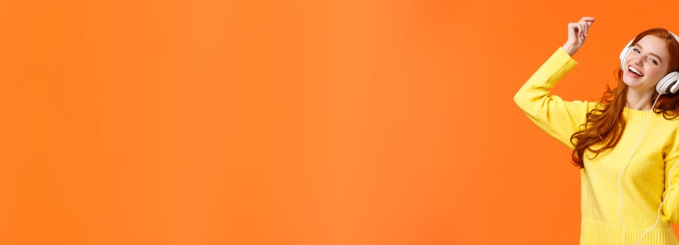 Carefree happy redhead teenage girl receive new headphones for christmas gift, dancing lift hand up while listen favorite music, holding smartphone, put awesome track, orange background photo