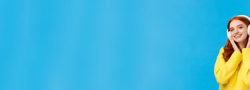 Fashionable attractive, carefree redhead woman in yellow sweater listening music with headphones, smiling tilting head joyfully, receive new earphones as christmas gift, standing blue background photo