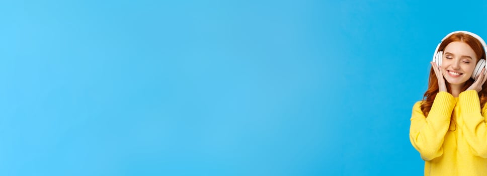 Technology, gadgets and lifestyle concept. Charming carefree redhead woman enjoy listening music in large white headphones, close eyes and smiling romantically, standing blue background photo