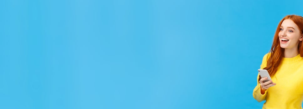 Cute and friendly, outgoing woman with red hair holding smartphone, messaging and waving at someone to turn attention, saying hi, smiling with hello, standing blue background photo