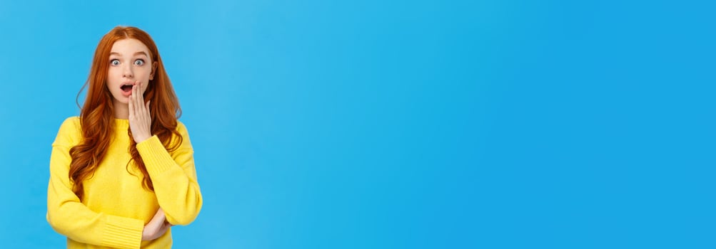 Wow stunning. Surprised girl found out secret or gossiping left shocked knowing truth. Astonished redhead woman stare camera speechless, gasping cover opened mouth amazed, blue background photo