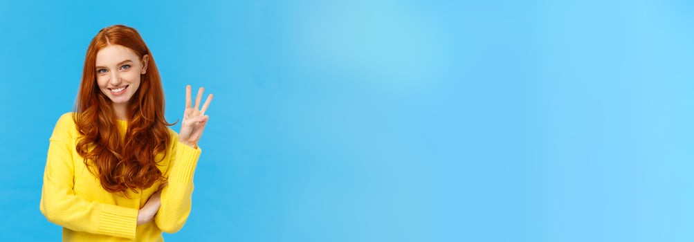 Gorgeous young redhead girl 20s in yellow sweater, smiling carefree making order, reservetion for three people, showing number third with fingers, standing blue background photo