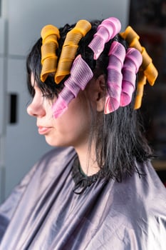 A woman sits in a salon chair with her hair in rollers, preparing for a perm photo