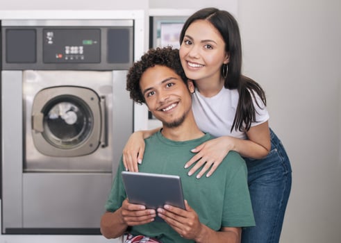 Laundry Service. Happy Young Diverse Couple Posing With Digital Tablet Hugging Near New Washing Machine Indoors, Booking Washer And Smiling To Camera Advertising Laundromat Service photo