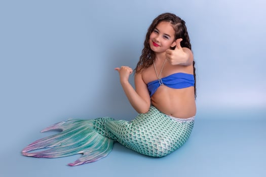 A happy girl in a mermaid costume sitting on a blue background showing thumbs up. photo