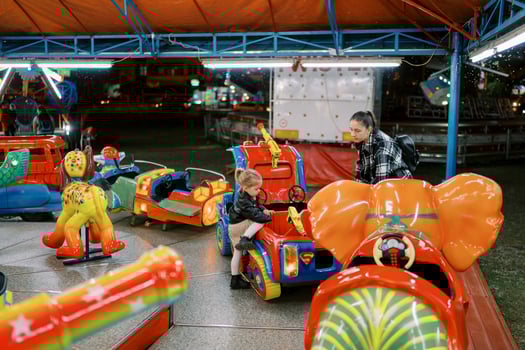 Mom stands near a toy car on a carousel into which a little girl climbs photo