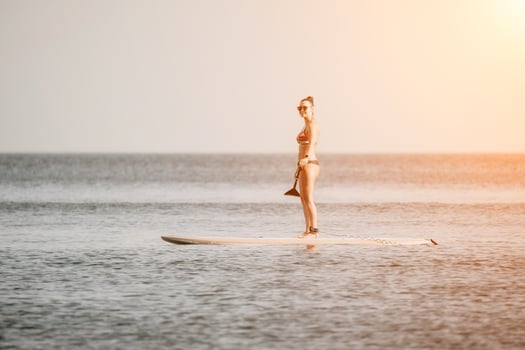 Sea woman sup. Happy woman in a rainbow-coloured bikini stands on SUP board. She is paddling through the calm water, creating ripples along the way. Idyllic sunset. Active lifestyle at sea or river. photo