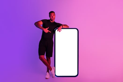 Young black fitness guy standing near huge smartphone with blank screen, pointing finger at device demonstrating mobile application for online workout over purple studio background, mockup photo