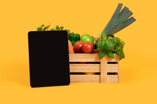 Digital Tablet With Blank Screen Standing Near Wooden Box With Fresh Vegetables And Fruits Over Yellow Studio Background. Mockup For Agriculture Website Advertisement Or Online Grocery Shopping photo