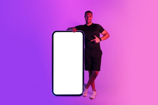 Muscular athletic african american guy leaning on huge phone, pointing at blank screen for application mockup, advertising mobile service or app for workout and sport on purple neon background photo