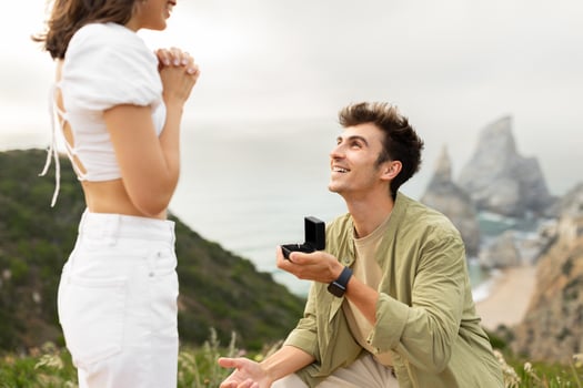 Happy young man standing on his knee and proposing to his girlfriend on a rocky cliff by the ocean shore, holding box with ring and looking at her beloved lady photo