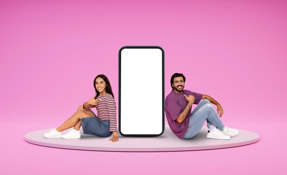 Relaxed positive happy millennial indian man and woman lovers chilling on platform next to huge phone with white empty screen, showing nice dating mobile app, pink background, mockup, blank space photo