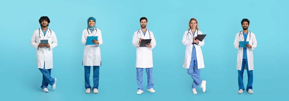 Smiling mature arabic and caucasian doctors in white coats with tablets and clipboards isolated on blue studio background, full length. Health care, exam, work, medicine and help photo