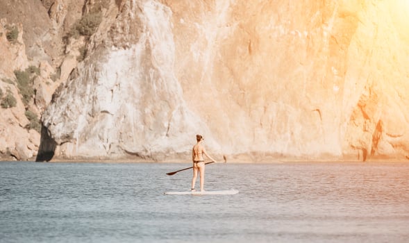 Sea woman sup. Silhouette of happy middle aged tanned woman in rainbow bikini, surfing on SUP board, confident paddling through water surface. Idyllic sunset. Active lifestyle at sea or river. photo
