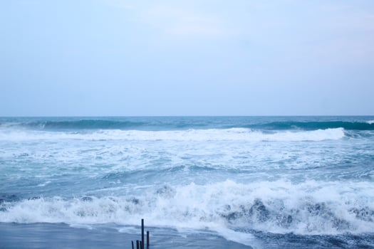 As the tide sweeps in at South Jogja Jetis Beach, Cornwall under a serene sky, waves gently roll, accompanied by ripples and bubbling foam. photo