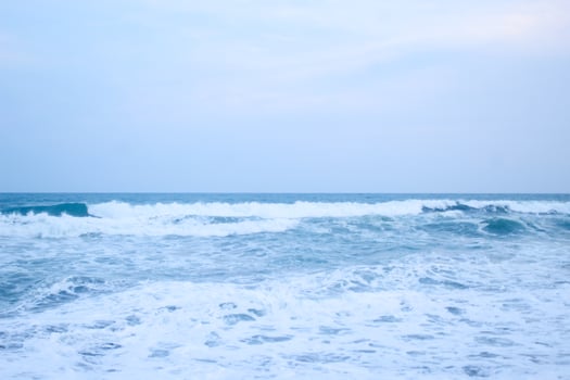 As the tide sweeps in at South Jogja Jetis Beach, Cornwall under a serene sky, waves gently roll, accompanied by ripples and bubbling foam. photo