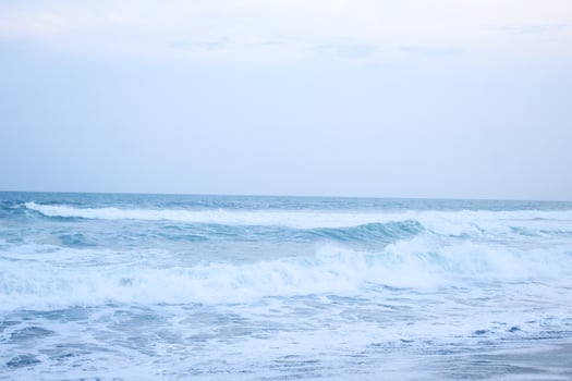 As the tide sweeps in at South Jogja Jetis Beach, Cornwall under a serene sky, waves gently roll, accompanied by ripples and bubbling foam. photo
