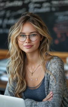 Smiling teacher wearing glasses sits at a desk with a laptop in front of a blackboard photo