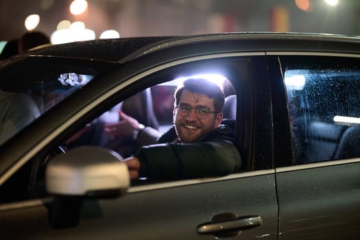 A modern businessman, donning eyeglasses, confidently drives his sleek car through the urban cityscape on a rainy night, epitomizing sophistication and success in his professional endeavors photo