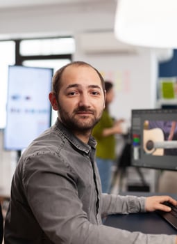 Man editor looking at camera, working with footage and sound on computer with two displays. Videographer editing movie smiling while processing film montage sitting in startup office. photo