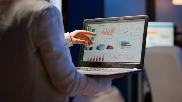 Back view of business woman analysing financial statistics holding laptop standing in start-up office late at night overworking. Busy focused employee using modern technology network wireless overtime photo