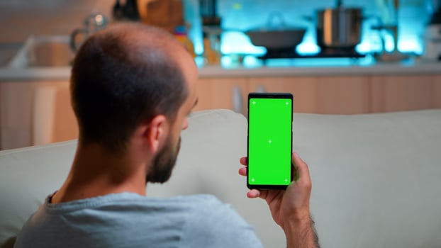 Caucasian male reading online social information on phone with mock up green screen chroma key display. Tired man sitting on sofa using isolated smartphone with technology internet photo