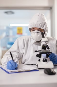 Doctor writing notes dressed in ppe suit and visor mask using microscope. Chemist in coverall working with various bacteria, tissue blood samples for antibiotics research. photo