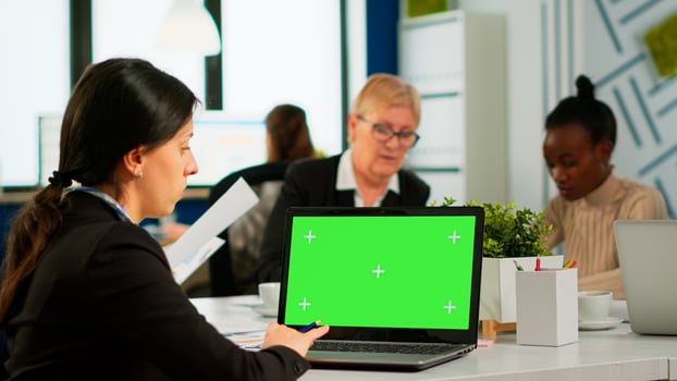 Over the sholder of manager sitting at meeting desk looking at laptop with green screen mockup while diverse team working on background. Multiethnic people planning project on chroma key display photo