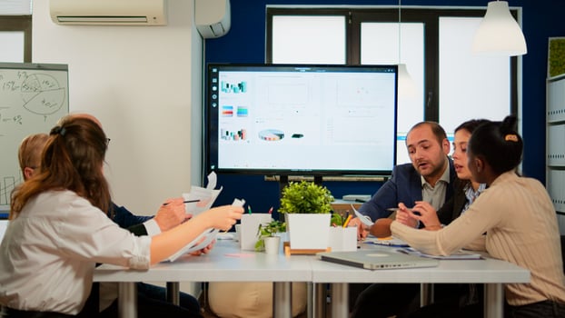 Executive explaining company's vision and potential to employees sitting at brainstorming table in broadroom with TV screen on the wall showing corporate growth photo