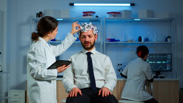 Young man wearing brainwave scanning headset discussing with neurological researcher about symptoms of disease. Medical scientist writing informations of brain scan on tablet working in laboratory photo