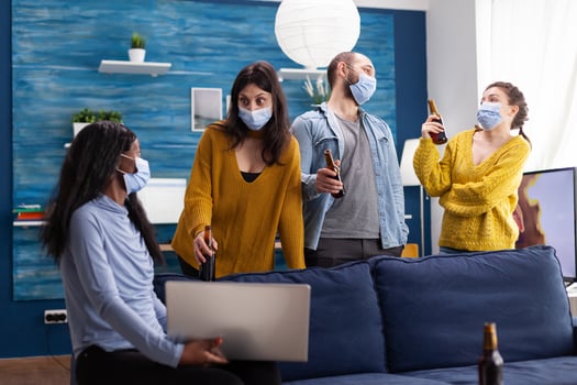 Multiracial group of friends chatting using laptop keeping social distancing and wearing face mask to prevent covid spread and infection in apartment living room. Conceptual image photo