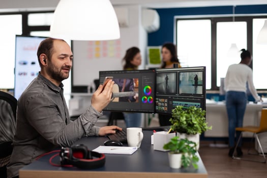 Multimedia creator artist talking on videocall with client holding smartphone editing footage sitting in creativity production agency. Videographer using post production software photo