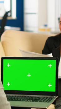 Over the sholder of manager sitting on couch talking with colleague holding laptop with green screen while diverse team working on background. Multiethnic people planning project on chroma key display photo