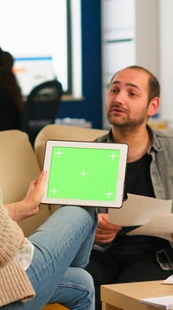Back view of businesswoman sitting on couch using tablet with green screen talking with colleague while team working on background. Multiethnic coworkers planning project on chroma key display photo