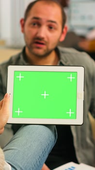 Over the sholder of manager woman sitting on couch talking with coworker holding tablet with green screen while diverse team working. Multiethnic people planning project on chroma key display photo