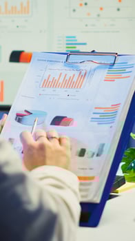 Close up of business woman holding clipboard with graphics and financial statistics working overtime in front of computer sitting in start-up business office. Busy employee using modern technology photo