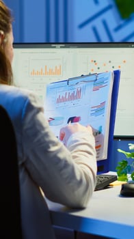 Back view of manager analysing graphics and financial statistics from clipboard working overtime in front of computer sitting in start-up business office. Busy focused employee using modern technology photo