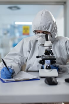 Biologist researcher wearing protective suit analyzing chemicals using medical microscope while writing clinical expertise during scientific experiment. Specialist doctor working in biochemistry lab photo