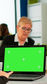 Over the sholder of manager sitting at meeting desk looking at laptop with green screen mockup while diverse team working on background. Multiethnic people planning project on chroma key display photo