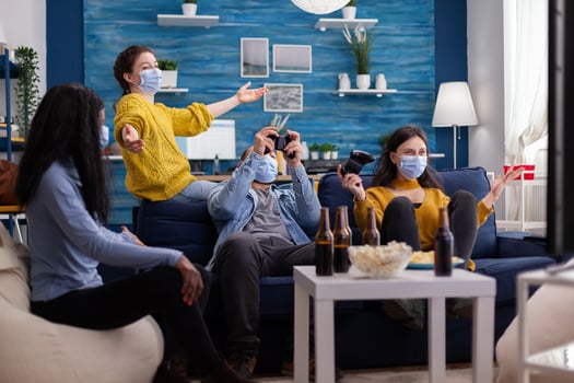 Cheerful group of multi ethnic friends celebrating victory against multi ethnic friends in home living room wearing face mask keeping social distancing during global outbreak with covid. New normal party. photo