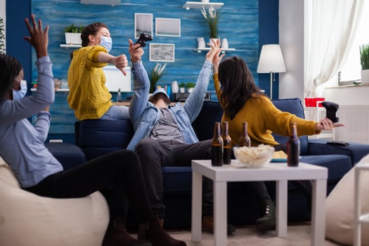 Mixed race friends celebrating games victory wearing face mask during coronavirus pandemic holding wireless joystick new normal party keeping social distancing. photo