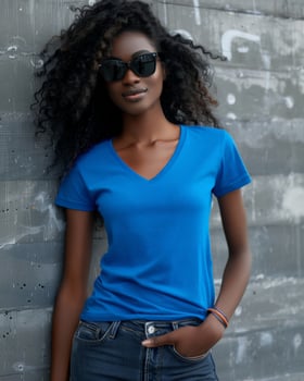 A young woman with curly hair wears a blue v-neck t-shirt, sunglasses, and jeans while standing by a wall photo
