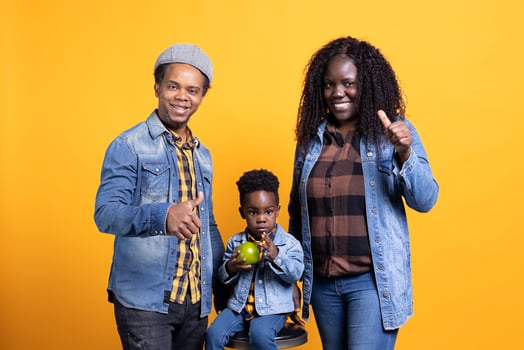 African american parents doing thumbs up symbol in studio, posing next to their cute little boy on the chair. Pleased grateful mother and father giving like gesture, agreeing on yellow background. photo