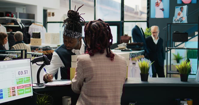 African american guy picking up order from clothing store, signing confirmation papers at cash register. Woman employee preparing boxes with requested fashion items and accessories. Camera B. photo