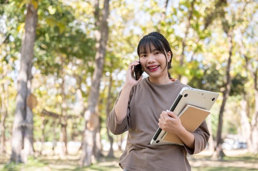 An Asian woman is talking with someone on her phone while walking in the park with her stuff. photo